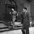 Milton H. Greene photographed this vintage Irish Fashion image in 1955 while on assignment for Life magazine. The focus is the model, standing on a step in front of an old brick building wearing a smart, calf-high button skirt. An older, crazy looking man in a suit is walking with a cane and reaching for a cigarette in his mouth to the right of the image. The model looks elegantly annoyed by the whole thing.