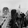 Marilyn Monroe in black and white photo by Milton H. Greene is seen walking off an American Airline plan in Los Angeles in 1956. Marilyn Monroe is wearing a sleek black dress, earrings and an dark fur coat over her back while she tilts her head back with a smile coming down the plane.