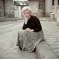 Taken on the 20th Century Fox backlot in 1954 by Milton H Greene, this color photograph of Marilyn Monroe shows her looking sad while sitting ton a curb and wearing a peasant dress. Her right hand is nestled under her chin.