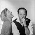 Eli Wallach has a serious look on his face as he stares directly at the camera with his left arm to his chin while his wife, Anne Jackson is seated to Eli's right and laughing. This black and white photograph was taken by Milton H Greene in his New York studio for Glamour magazine in 1964.