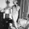 Black and white photograph by Milton H. Greene of barefoot Marilyn Monroe inside of Regis Hotel standing on an couch wearing white pants and an rolled up white shirt while answering a phone with one hand balanced on a ornate table top.