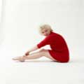 Wearing nothing but a red sweater, Marilyn Monroe is seated with her legs out in front of her with her hand touching her ankles. Looking directly at the camera she is smiling against a white background. Milton H Greene took this photo in 1955.